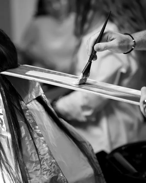 Mit einem schmalen Pinsel wird Farbe auf eine Haarsträhne gestrichen, die auf einem Brett liegt; darunter hängen Alufolien.