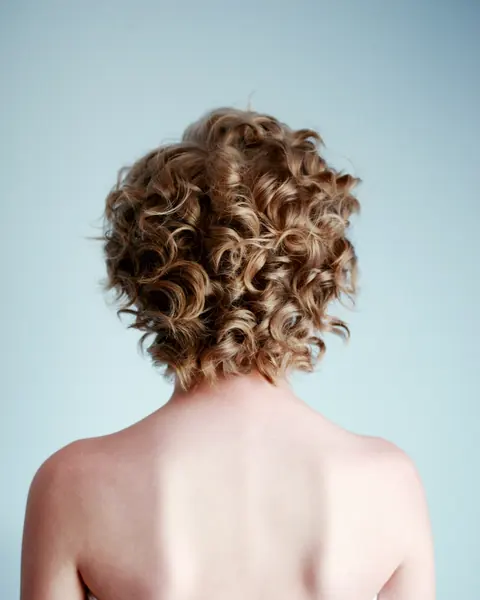Kurze, blonde Korkenzieherlocken von hinten, die Schultern sind frei.