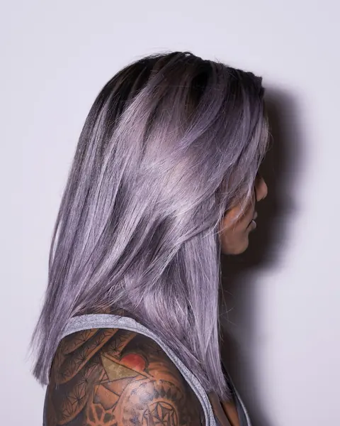 Lange, glatte Haare in Lavendel mit dunklem Ansatz, seitlich von hinten, darunter eine tätowierte Schulter.