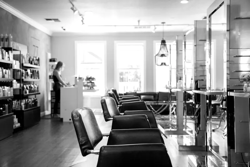 Salonraum mit einer Reihe schwarzer Friseurstühle vor hohen Spiegeln, hinten große Fenster und ein Regal mit Pflegeprodukten.