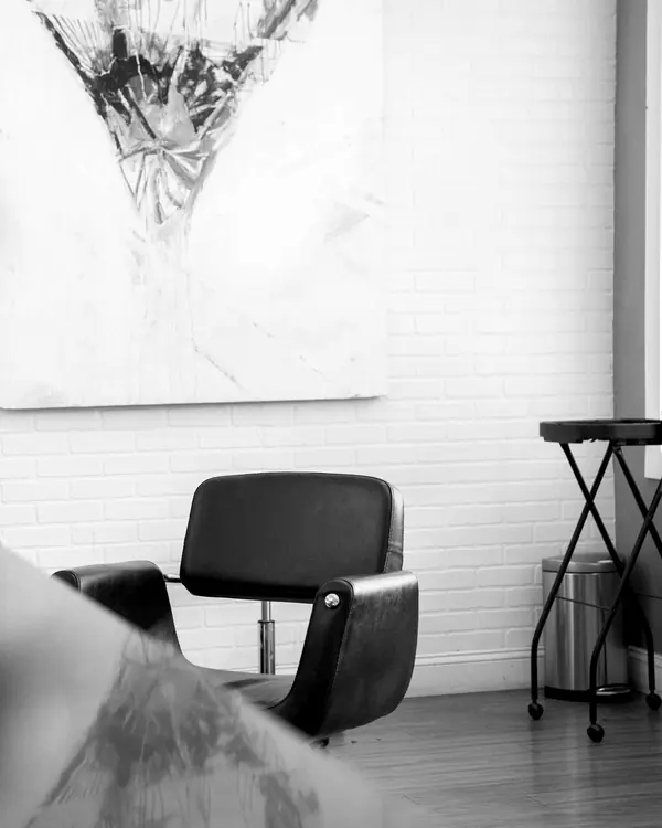Ein einzelner schwarzer Friseurstuhl vor einer weiß gestrichenen Backsteinwand, darüber ein großes abstraktes Bild.